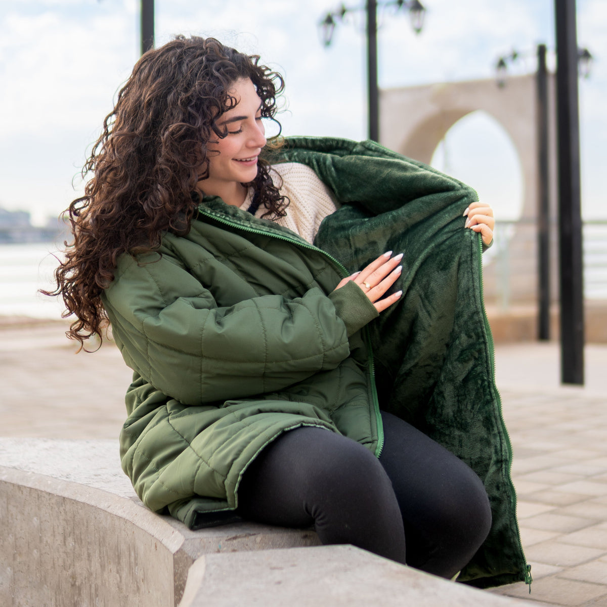 Woman sitting on a bench wearing a green puffer jacket with a hood, holding the hood over her head.
